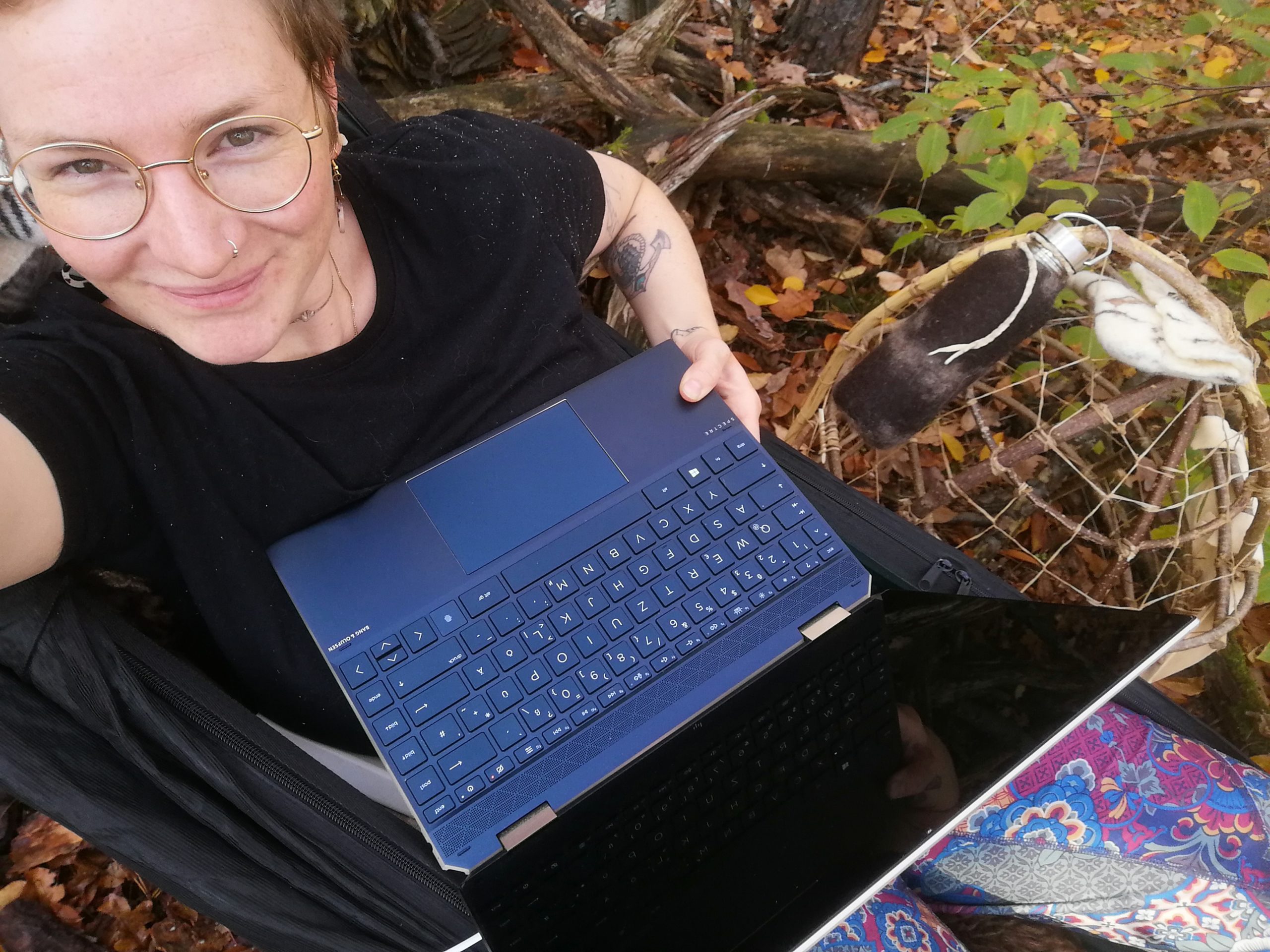 Elena liegt mit Laptop in der Hängematte im bunt belaubten Herbstwald. Neben ihr ist ein Packkorb aus Holz zu sehen.
